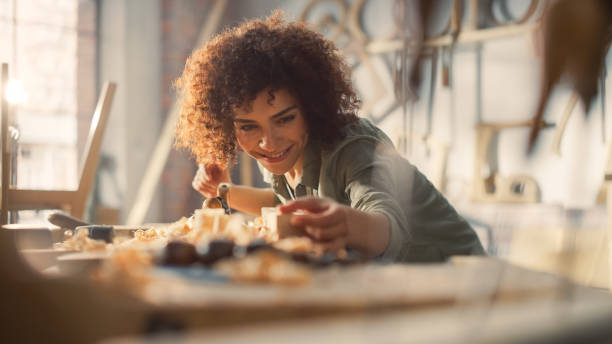 close up portrait of an attractive young artisan carpenter using hand plane to shape a wood bar. multiethnic female working on a project in a loft studio with tools on walls. - carpenter restoring furniture wood imagens e fotografias de stock