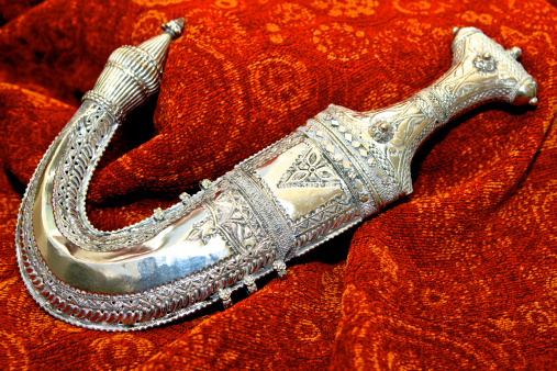 Arabian Dagger with Silver hilt, in a silver sheath on rich red background. It is called a Khanjar in Oman and the UAE; a Jambiya in the Yemen.  This one is a Jambiya.  There are many uses for the dagger. It is a symbolic weapon, and worn by men after puberty. These days it is used as part of formal dress, quite often on ceremonial occassions, to go with the Arabian robe. Many Sheikhs of the Arabian Gulf wore such daggers on ceremonial occasions, in the past. The unsheathing of the dagger was considered a social taboo. Men would only do that in a situation of conflict, if they sought revenge or assassination. Photo shot in natural light; horizontal format. Copy space.