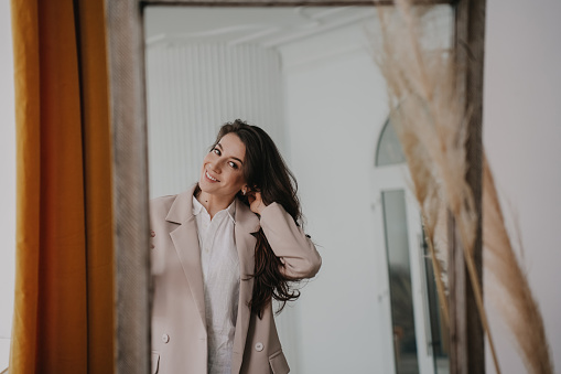 Cheerful brunette hispanic woman at mirror trying on beige coat at fitting room. Attractive calm caucasian model with long loose hair prepares for date. Women beauty. Hairstyle. Businesswoman at home.