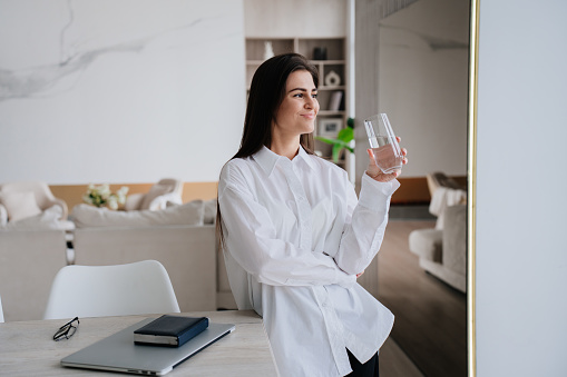 Pretty caucasian brunette girl in white shirt holds glass of water leans on desk with laptop, diary, glasses looks away dreamily standing at living room. Successful hispanic businesswoman relaxing.