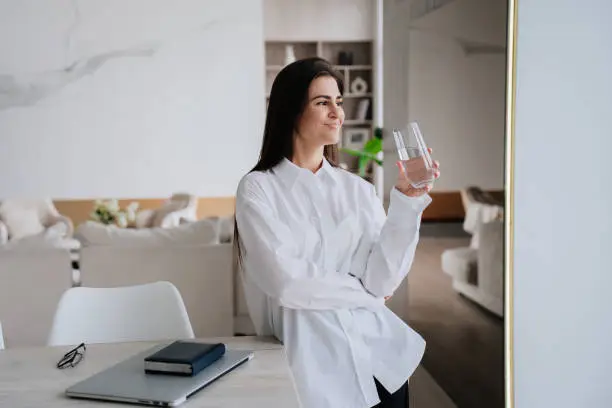 Photo of Pretty caucasian brunette girl in white shirt holds glass of water leans on desk with laptop, diary, glasses looks away dreamily standing at living room. Successful hispanic businesswoman relaxing.