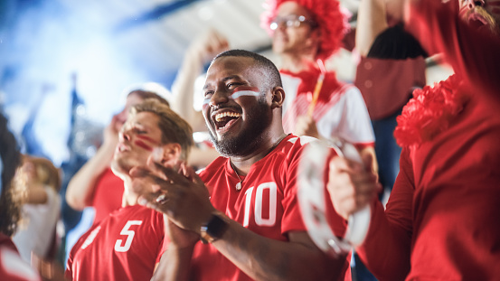 Sport Stadium Soccer Match: Diverse Crowd of Fans Cheer for their Red Team to Win. People Celebrate Scoring a Goal, Championship Victory. Group People with Painted Faces Cheer, Shout, Have Fun