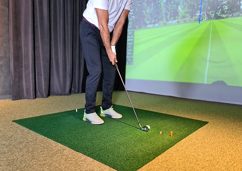 Golf player playing golf indoors on golf simulator. Driving range with screen for golf and golfer legs