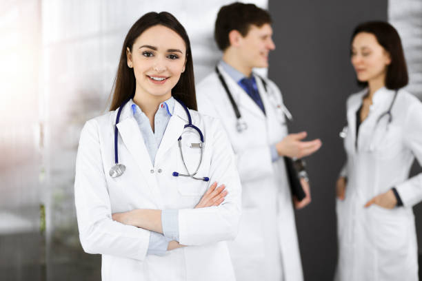 Smiling intelligent woman-doctor is standing with arms crossed in sunny clinic together with her colleagues at the background. Perfect medical service in a hospital Smiling intelligent woman-doctor is standing with arms crossed in sunny clinic together with her colleagues at the background. Perfect medical service in a hospital. Suboxone Doctors stock pictures, royalty-free photos & images