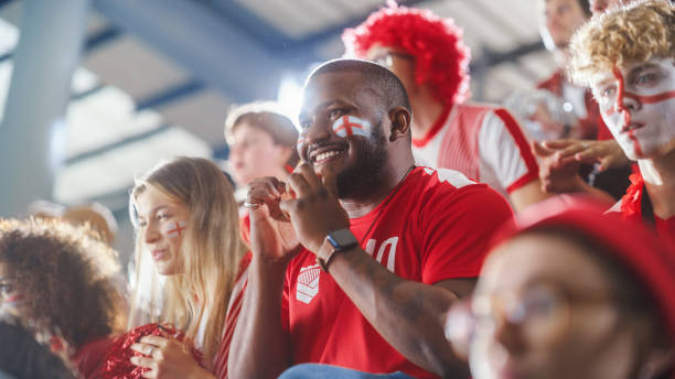 istock Sport Stadium Big Event: Handsome Expressive Black Man with Painted face Jumping Up and Cheering Goal Score. Crowd of Fans Shout for Red Soccer Team to Win. People Celebrate Championship Victory 1479689319