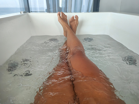 Close-up POV shot of unrecognizable legs crossed at ankle inside a bathtub with water jets flowing from the side