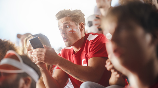 Sport Stadium Soccer Match: Caucasian Man Using Smartphone Cheering for Red Team to Win, Looking at Mobile Phone to Check App, Bet, Score, Winnings. Crowd Celebrate Goal, Championship Victory