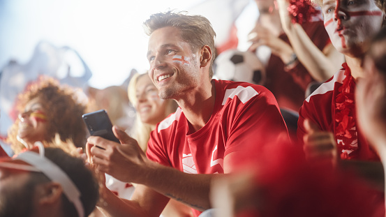 Sport Stadium Soccer Match: Caucasian Man Using Smartphone Cheering for Red Team to Win, Looking at Mobile Phone to Check App, Bet, Score, Winnings. Crowd Celebrate Goal, Championship Victory