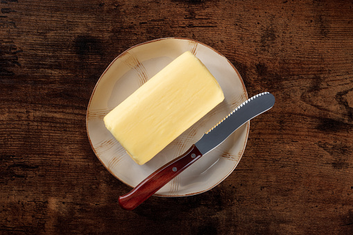 A block of butter with a knife on a plate, overhead flat lay shot on a dark rustic wooden background