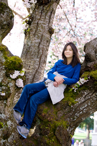 Young biracial teen girl sitting in cherry tree in full bloom