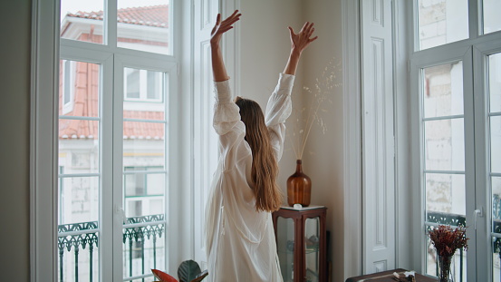 Relaxed girl stretching arms at window interior closeup. Calm lady greeting new day enjoying weekend at cozy apartment. Dreaming woman waking up resting at domestic place back view. Morning concept