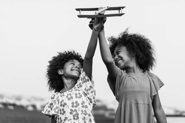 irmãs gêmeas afro correndo na praia enquanto brincam com avião de madeira - foco nas mãos segurando brinquedo - edição em preto e branco - airplane black and white fun child - fotografias e filmes do acervo