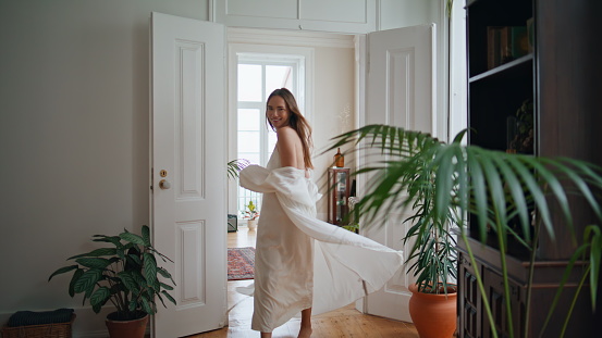 Active woman spinning at home interior. Smiling lady in silk pyjamas having fun dancing at light apartment. Positive girl moving alone at cozy house with plants. Excited female person circling at hall