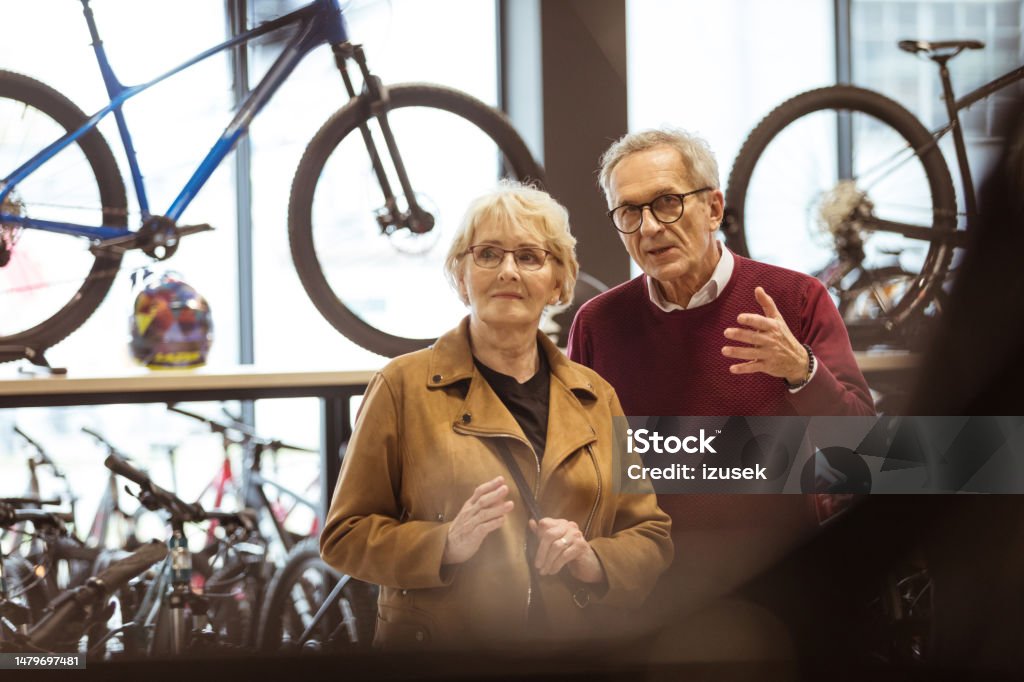 Senior couple in bicycle shop Senior woman and senior man watching bicycles in bike shop. Man pointing with index finger. Electric Bicycle Stock Photo