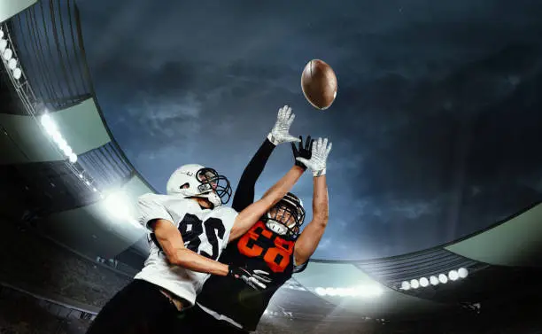 Dynamic shot of male sportsmen, professional american football players during game, catching ball. Open air 3D stadium in evening. Concept of sport, competition, action and motion, game, cup.