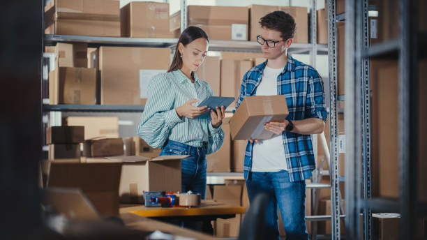 istock Female Inventory Manager Shows Information on Tablet Computer to a Worker Holding a Cardboard Box, They Talk and Discuss Work. Stock of Parcels with Products Ready for Shipment in the Background. 1479701794