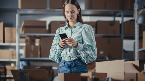 Portrait of a Beautiful Small Business Owner Using Smartphone at Work in a Warehouse. Female Connecting with Colleagues, Supporting Clients Over Internet. Sales Manager Working in Online Store. retrato de una hermosa propietaria de una pequeña empresa que usa un teléfono inteligente en el trabajo en un almacén. mujer conectándose con colegas, apoyando a clientes a través de internet. gerente de ventas trabajando en tienda online.