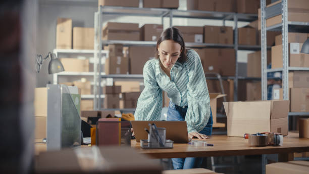 Small Business Owner of an Online Store Works on Laptop Computer while Standing at Her Desk in Warehouse. Female Employee Packing a Stylish Yellow Jumper in the Room with Shelves Full of Parcels. la propietaria de una pequeña empresa de una tienda en línea trabaja en una computadora portátil mientras está de pie en su escritorio en el almacén. empleada empacando un elegante jersey amarillo en la habitación con estantes llenos de paquetes.