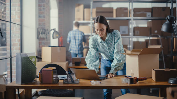 istock Small Business Owner Works on Laptop Computer while Standing at Her Desk in Warehouse. Female Employee Packing an Online Order in the Room with Shelves Full of Cardboard Box Parcels. 1479704657
