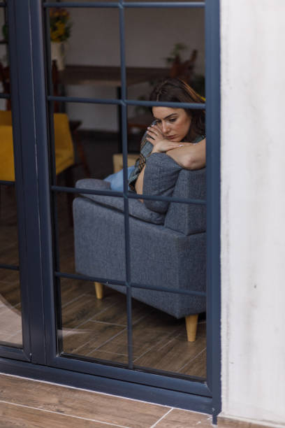 Portrait of sad young woman sitting on the sofa in her living room, wrapped in a blanket, leaning against the backrest, hugging self, looking down, contemplating. Seen through glass window.