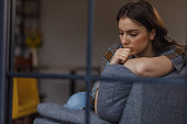 Sad young woman sitting on the sofa, wrapped in a blanket, contemplating problems and solutions