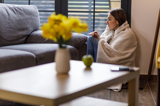 Wide shot of distraught young woman sitting on the floor, by the window, wrapped in a cozy blanket, trying to comfort herself when battling negative emotions. Focus on background.