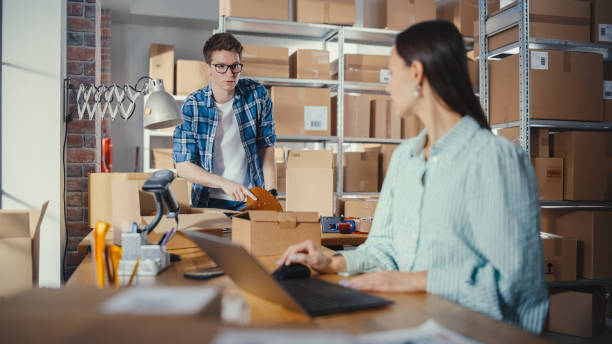 istock Young Male and Female Warehouse Inventory Managers Packing Orders for Clients, Using Laptop Computer and Checking Retail Stock. Employee Preparing a Parcel with Stylish Retro Bicycle Seat. 1479709811