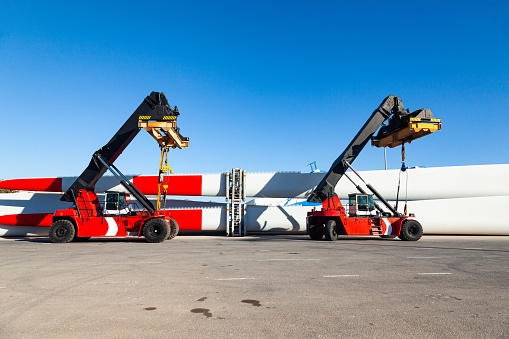 A four-wheeled mobile container loaders parked at the seaport, next to the storage blades for wind turbines.