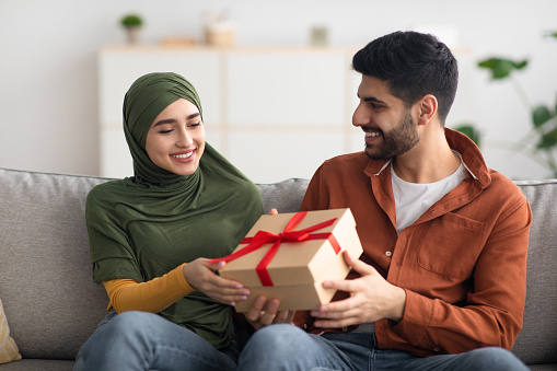 Happy Muslim Man Giving Gift Box To Wife Congratulating Her Sitting On Sofa At Home. Couple Celebrating Family Holiday. Birthday Present And Celebration Concept