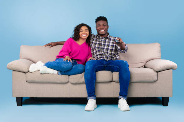 istock Happy young black couple with remote control sitting on sofa and watching TV over blue studio background, full length 1479720873