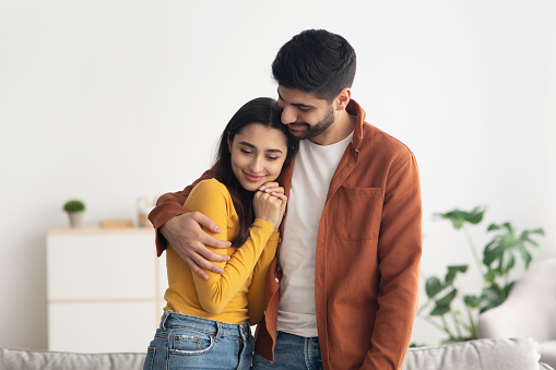 Happy Middle Eastern Couple Embracing Standing At Home. Young Arabic Husband Hugging His Beautiful Wife Indoor. Love And Romantic Relationship. Successful Marriage Concept