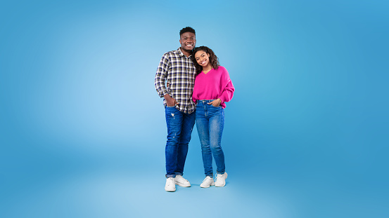 Full length portrait of happy young African American couple in casual wear hugging and looking at camera on blue studio background, panorama. Cheerful millennial spouses posing and smiling together