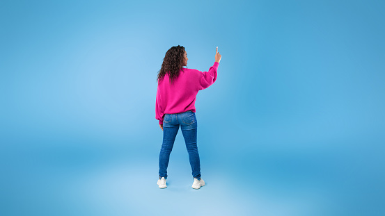 Back view of millennial African American lady touching imaginary screen, pressing button, using virtual interface on blue background, banner design with free space. Contemporary technologies concept