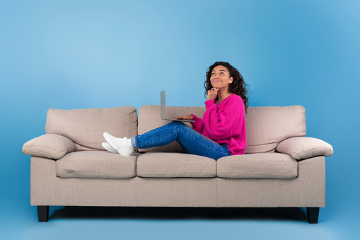 Thoughtful young black woman in casual wear working online, sitting on couch and using laptop on blue studio background. Cheerful African American lady surfing internet on portable pc