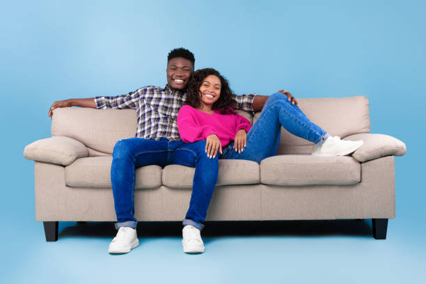 istock Happy millennial black couple relaxing on sofa, smiling at camera on blue studio background, full length 1479721836