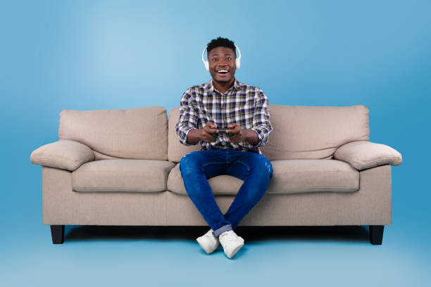 istock Happy young black guy with joystick playing video game, wearing headset, sitting on couch over blue studio background 1479721840