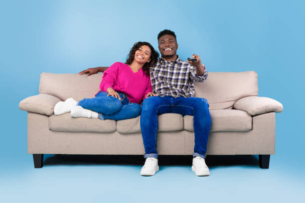 青いスタジオの背景にリモコンを持つ興奮した若い黒人夫婦がソファに座り、テレビを見ている - home interior entertainment sofa men ストックフォトと画像