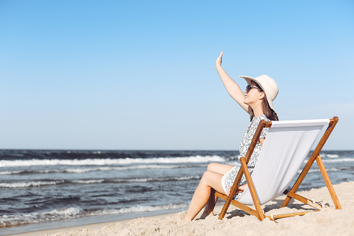 Happy brunette woman sitting on a wooden deck chair at the ocean beach while waving and greeting somebody with her hand.