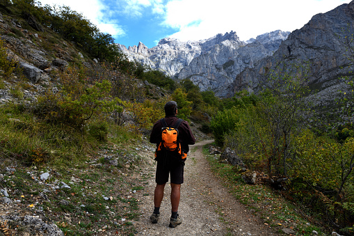 The hiker is in the middle of the path looking up at the mountains. The path is gravel and leads towards mountains. There are bushes either side. Towering about the path are the rocky mountains of the Picos de Europa National Park. The sky is blue at the top of the photo, with a few clouds. 

The photo is taken at Posada de Valdeón, in the province of León, Castile and León, Spain. It is in the Picos de Europa National Park