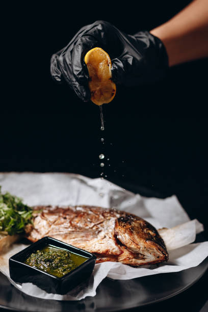 Chef squeezes lemon juice on golden fried fish on a black plate covered with parchment, decorated with pesto sauce and lemon.