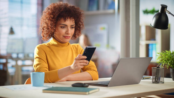 istock Beautiful Middle Eastern Manager Sitting at a Desk in Creative Office, Using Smartphone. Young Stylish Female Sending Message, Browsing Internet. Colleagues Working in the Background. 1479739442