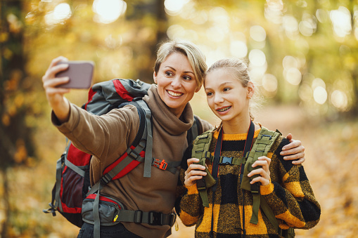 Shot of a teen girl and her mom taking a selfie with smartphone during walk together through the forest in autumn.