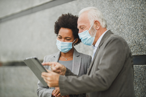 Shot of successful black businesswoman and her senior male colleague with protective mask using digital tablet and having a discussion while standing against a wall of corporate building during COVID-19 pandemic.