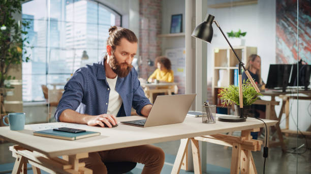 istock Creative Project Manager Sitting at a Desk, Working on a Laptop Computer in an Office. Boho Style Inspired Handsome Marketing Specialist Planning a Project, Browsing Internet and Writing Emails. 1479740703
