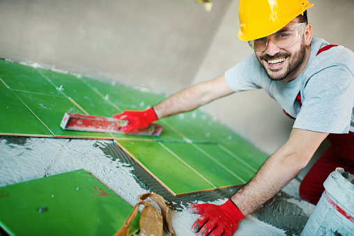 Closeup side view of a handyman installing green ceramic tiles over apartment floor.  He's wearing red gloves and a yellow helmet.