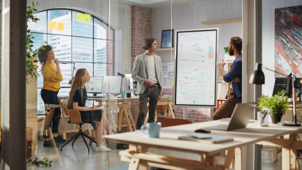 istock Junior Marketing Specialist Making a Team Presentation in Creative Office Meeting Room. Project Manager Showing a Fresh Product Timeline. Project Plan Presentation on Digital Whiteboard Monitor 1479741735