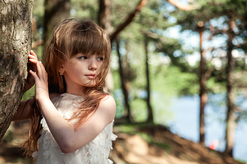 Pensive little girl in white dress standing near tree in greenery forest outdoors, looking away. Portrait of sad girl posing in sunshine woodland. Environmental protection concept. Copy ad text space