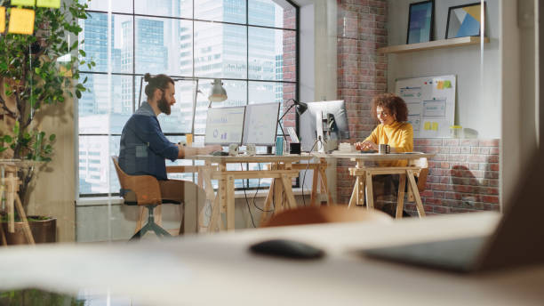 Two Specialists Sitting Behind Desks Next to Big Window with Downtown City View and Work with Reports on a Computer. Diverse Male and Excited Female Busy Researching and Analyzing Business Data. dos especialistas sentados detrás de escritorios junto a una gran ventana con vista al centro de la ciudad y trabajan con informes en una computadora. hombre diverso y mujer emocionada ocupada investigando y analizando datos comerciales.