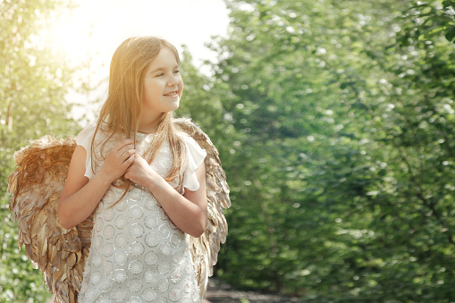 Smiling little girl in angel image with gold wings posing in greenery forest, happy looking away. Funny young lady in white dress in sunshine woodland. Fairytale, fantasy concept. Copy ad text space