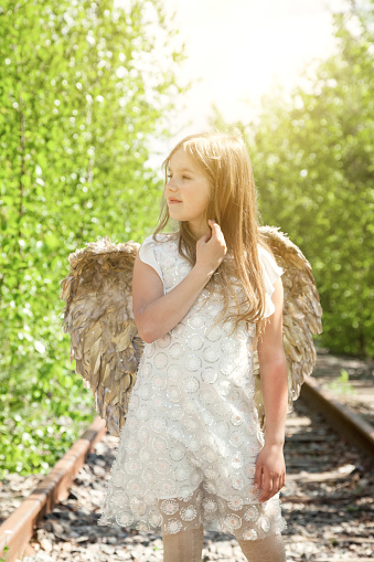 Pensive little girl in angel image with gold wings posing in greenery forest, thought looking away. Funny young lady in white dress in sunshine woodland. Fairytale, fantasy concept. Copy ad text space